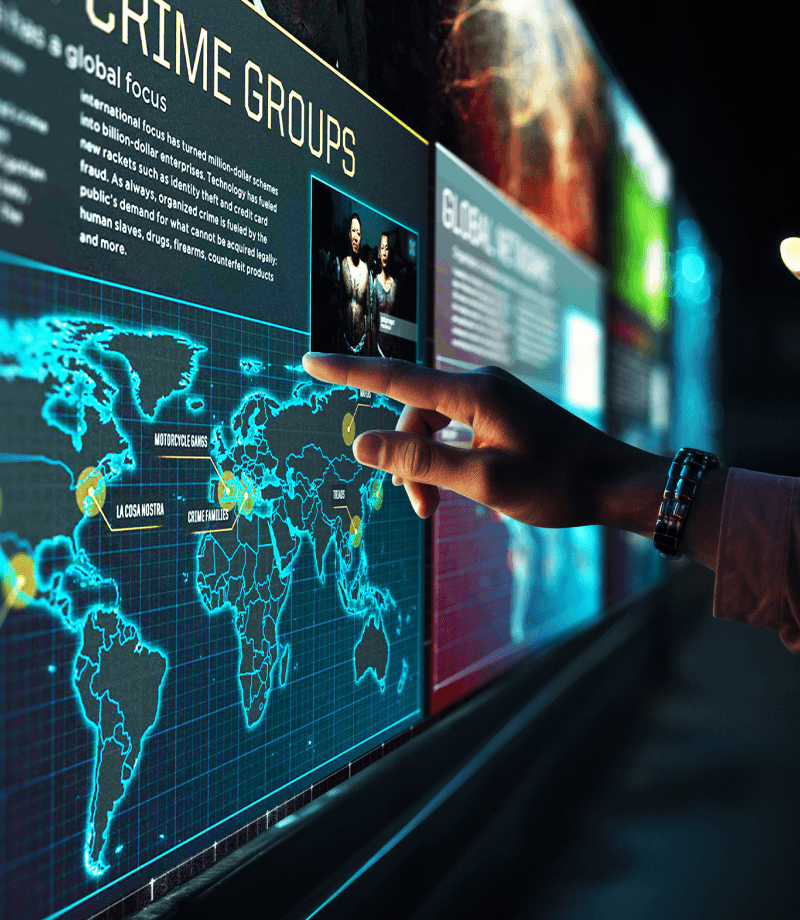 A close-up view of a museum visitor's hand with the index finger extended to touch a video screen, which is part of a large array of screens on a wall partially visible in the background. The screen displays part of a header text reading Crime Groups, above a world map with several cities highlighted in yellow.
